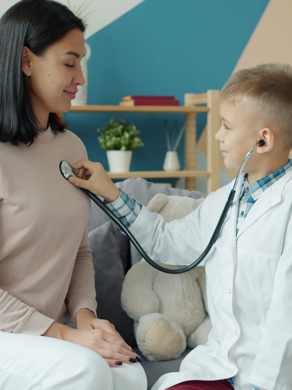 A little boy in a doctor’s coat listens to his mom’s heartbeat with a stethoscope